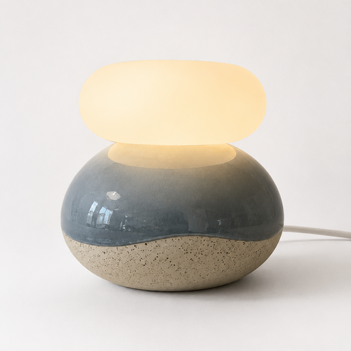 A modern ceramic table lamp with a rounded, dual-texture base in blue-gray and beige, topped with a smooth, oval-shaped frosted white shade that glows warm yellow when lit. 