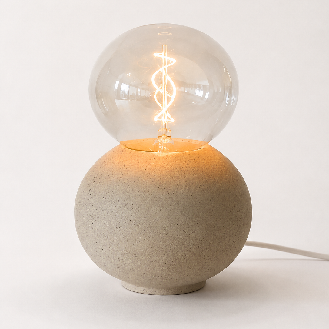 A modern table lamp with a matte concrete sphere base and a clear glass globe bulb on top, illuminated with a glowing spiral filament. 
