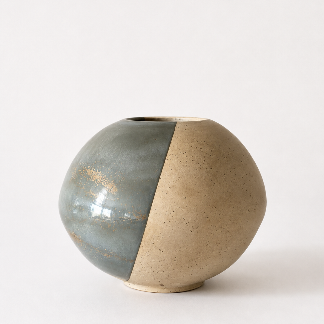 A spherical ceramic vase with a split design, featuring a matte speckled beige half and a glossy slate-blue half with subtle gold leaf flecks.