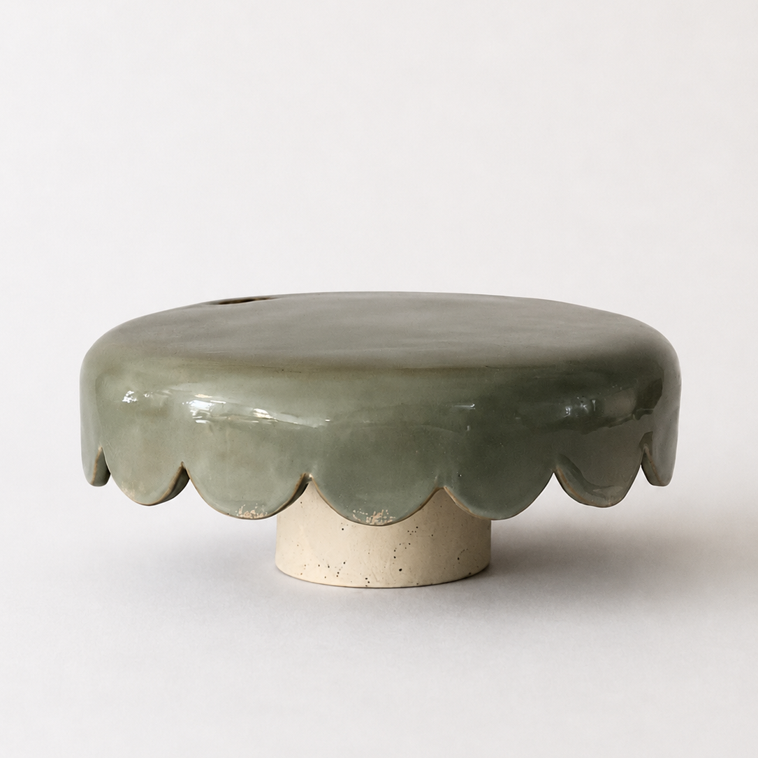 A hand-built ceramic pedestal piece with a soft, rounded silhouette and scalloped edge that gives it its slightly playful character. Finished in a muted grey-green glaze over a natural speckled clay base.